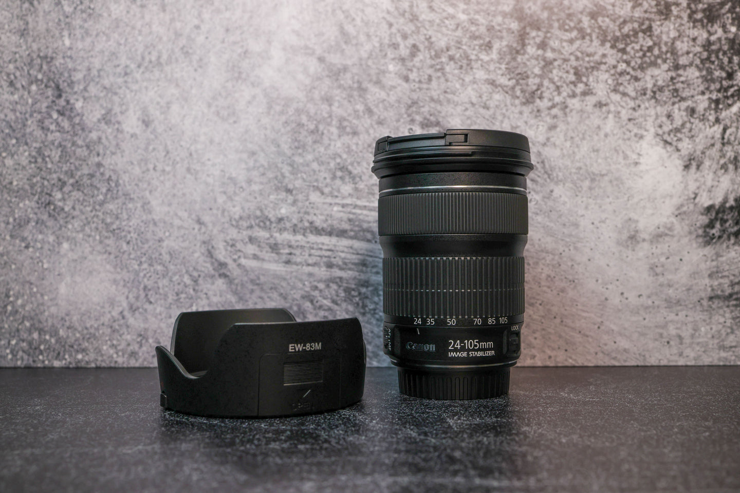 A Canon 25-105mm lens sitting vertically on the rear lens cap with the lens hood to the left side sitting on the surface.