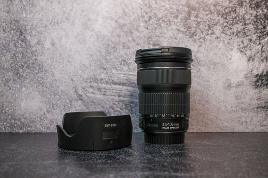 A Canon 25-105mm lens sitting vertically on the rear lens cap with the lens hood to the left side sitting on the surface.
