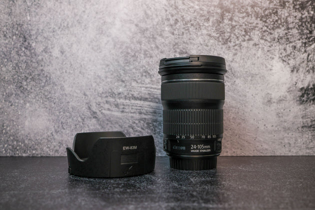 A Canon 25-105mm lens sitting vertically on the rear lens cap with the lens hood to the left side sitting on the surface.