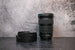 A Canon 25-105mm lens sitting vertically on the rear lens cap with the lens hood to the left side sitting on the surface.